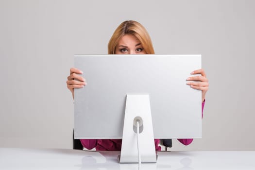 young woman sitting in the table and using computer on gray background. Surprised and scared hidden behind the monitor. It peeping from behind him