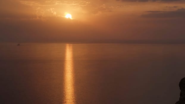 Breathtaking sunset over the Atlantic ocean with the bright golden sun coming down to the horizon. Aerial of the calm ocean, black cliff, and bronze sunset.