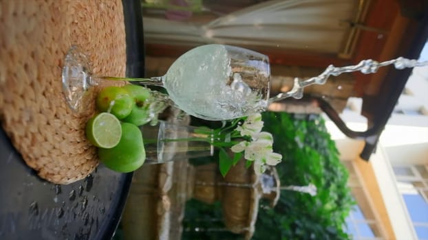 Refreshing drink is poured on summer veranda. Clip. Vertical shooting of illuminating streams of drinks in glass glasses. Pouring fresh drink with splashes in summer.