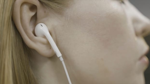Close up side view of blonde woman head wearing headphones and touching her hair. Girl with white earphones tucking her hair behind her ear.