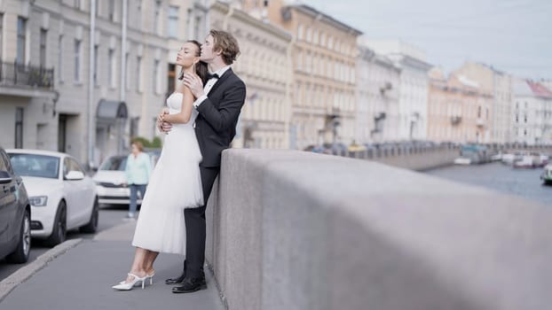 Side view of caring lovely just married couple embracing by the granite sidewalk. Action. Elegant bride and groom outdoors in the street