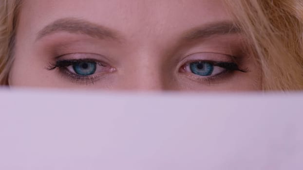 Closeup of woman face behind book cover. Close up of redhead girl with blue eyes behind book. Learning and reading concept. Women's eyes read close up.