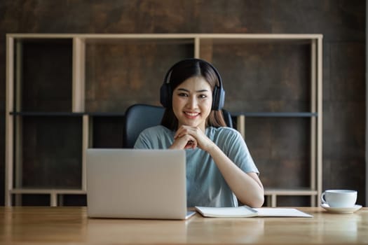 Young female student wearing headphones sits intently and happily studying online on her laptop in the living room at home..
