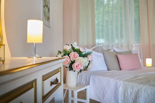 Cozy bedroom with bed located against mirror under dressing-table with lamp and window with curtain