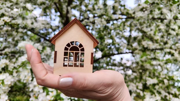 Small wooden toy house on palm of woman hand on natural background with apple flowers. symbol and concept of care, buying, selling, donating of eco friendly home. close-up. soft selective focus