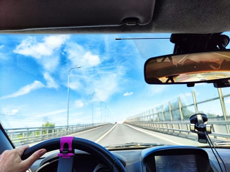 Car salon, steering wheel, hand of woman, view on nature landscape. View from seat of driver on Road, forest, blue sky, white clouds at sunny day. Concept of single trip of female traveller