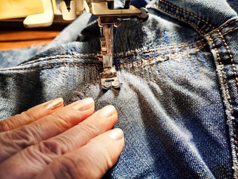 Sewing machine and blue jeans fabric. A woman's hand and fingers next to a sewing machine needle when sewing up tear in denim