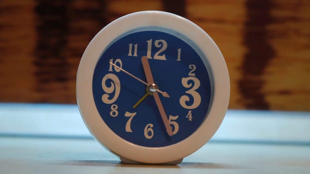 Close up of unusual alarm clock of blue color with white frame. Blue alarm clock with white numbers and metal hands standing on the table in the room.