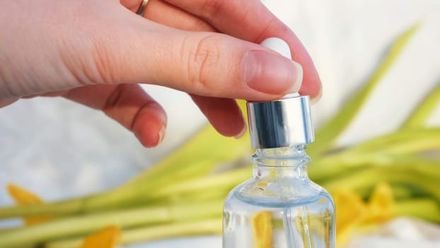 Vial with essential oil and yellow soft petals isolated on a white textile background. Girl hand touching cosmetic package surrounded by tulip leaves.