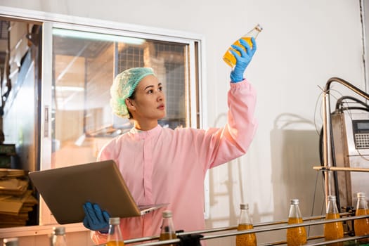 Woman worker in a beverage factory examines bottles on a conveyor belt employing a laptop for quality assurance ensuring high liquid standards.