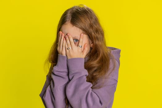 Portrait of curious young preteen child girl kid closing eyes with hand and spying through fingers, hiding peeping, exploring way, seeking something in distance. Children isolated on yellow background