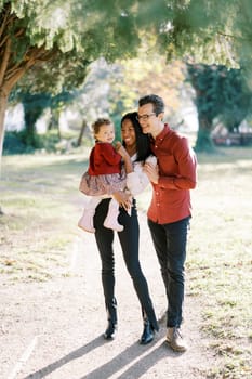 Dad hugs mom with a little girl in her arms while standing under a green tree. High quality photo