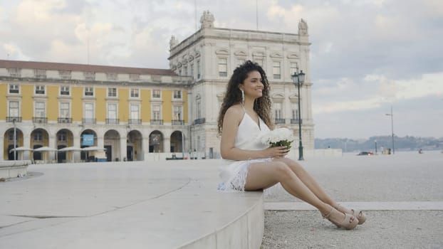 Beautiful young woman with bouquet of white flowers on street. Action. Attractive young woman smiles and holds bouquet of flowers. Smiling woman in dress and bouquet of flowers is waiting for date.