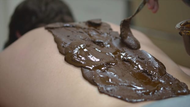 Massage with mud on back. Close-up massage therapist of Spa salon smears brown mixture on male back. Spa care, relaxation and treatment.