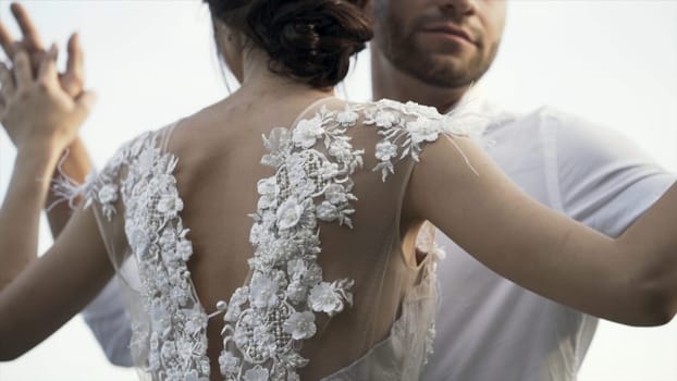 Rear view of a beautiful white wedding dress of a young slim bride with gentle flowers and cutout back. Loving ang hugging bride and groom on bright sky background.