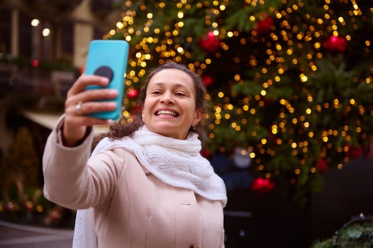 Pretty brunette woman smiling a toothy smile, holding smart mobile phone in her outstretched hand, making selfie and sharing her happy moments on social media during Christmas traditional market