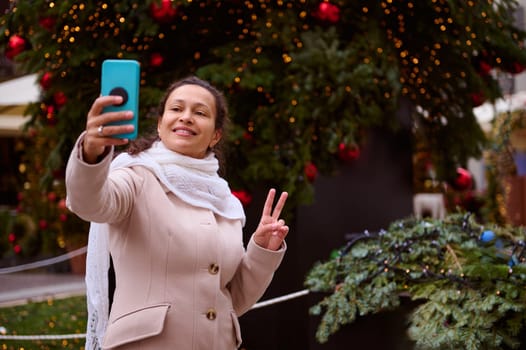 Beautiful woman in warm beige coat, standing against a Christmas tree decorated with garlands, shows peace sign while makes selfie and shares her lifestyle on social media during Christmas funfair