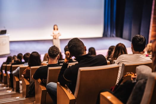 Business and entrepreneurship symposium. Female speaker giving a talk at business meeting. Audience in conference hall. Rear view of unrecognized participant in audience.