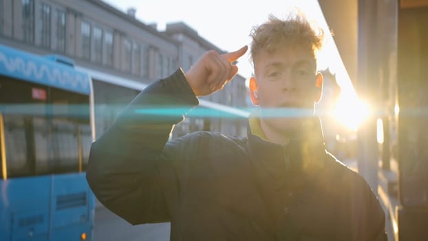 Cheerful young man dancing on street. Action. Young man is dancing merrily at bus stop on sunny day. Man in jacket and headphones dances while waiting for bus.