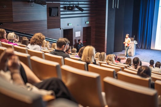 Business and entrepreneurship symposium. Female speaker giving a talk at business meeting. Audience in conference hall. Rear view of unrecognized participant in audience.