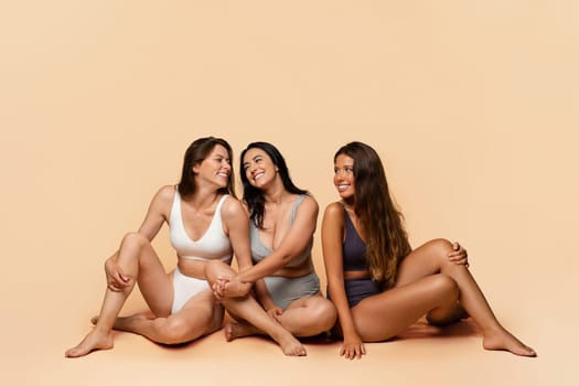 Three glad diverse millennial women in undergarments sit on floor, enjoy body positivity, beauty care, have fun, isolated on light studio background, full length. Friendship, confidence
