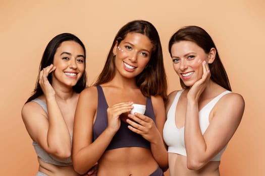 Three multiethnic young women in underwear show jar, put cream on face, enjoy beauty care, hydration at home on beige studio background. Fun lifestyle, cosmetics and spa, close up