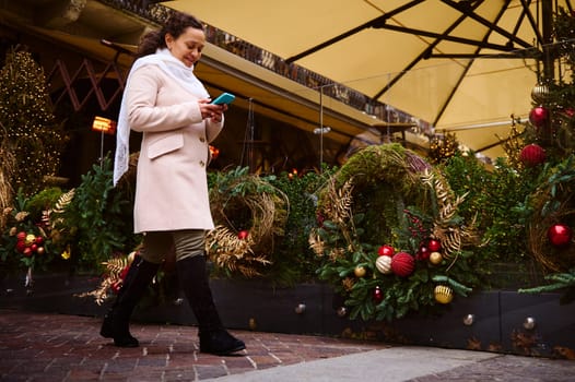 Full length portrait of a smiling authentic woman in warm beige coat, talking on a mobile phone, while walking on a city street decorated with festive garlands for celebrating New Year and Christmas