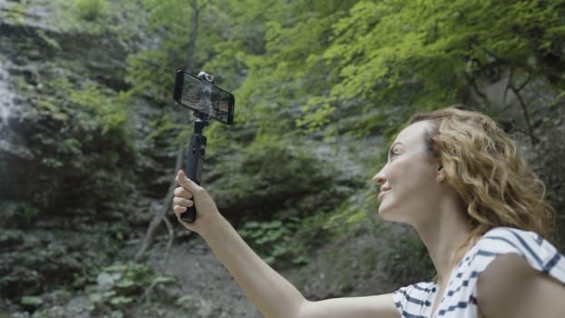 Woman taking pictures or shooting video of mountains and waterfall on her smartphone using a tripod. Action. Female traveler with a selfie stick and a phone standing in a gorge surrounded by green cliff slopes and waterfall