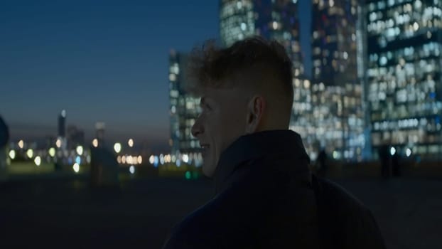 Man looks at modern glowing skyscrapers at night. Action. Young man on background of bright glowing skyscrapers at night. Man looks at skyscrapers glowing at night in city.