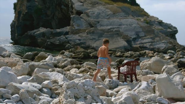 Boy sits on chair by sea. Creative. Boy walks alone on rocky shore with rocks. Boy sits on chair between rocks on shore on sunny day.