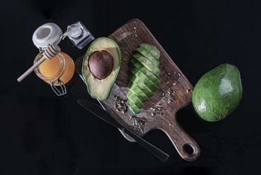 Fresh organic sliced avocado and Sliced avocado halves with spices on a wooden cutting board served with Honey and Salt on dark background. Healthy food concept, Space for text, Selective Focus.