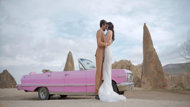 Beautiful bride and groom in wedding dress and suit near pink vintage cabriolet car. Action. Wedding couple standing near a convertible on a natural background