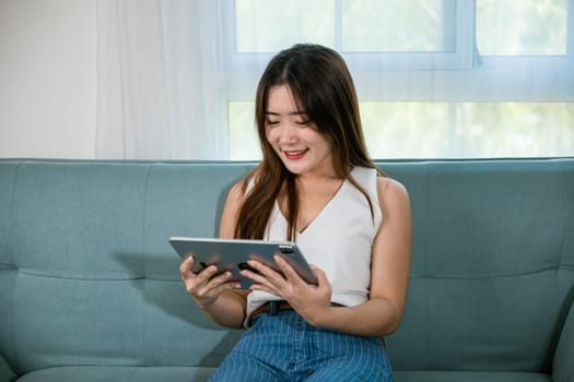 Happy Asian woman playing smart tablet at home while relaxing on sofa living room, Beautiful female sitting on couch using digital tablet pc, technology social media