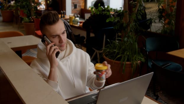 Working environment. Stock footage. A young man sitting in a cafe and working at a laptop while talking on the phone. High quality 4k footage