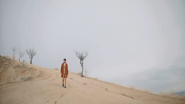 Man walking on desert hill. Action. Sand covered hill slope with bald trees and dry grass on cloudy sky background
