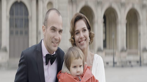 Man in suit, elegant woman in white dress, and a little girl in red jacket. Action. Happy family in a city square