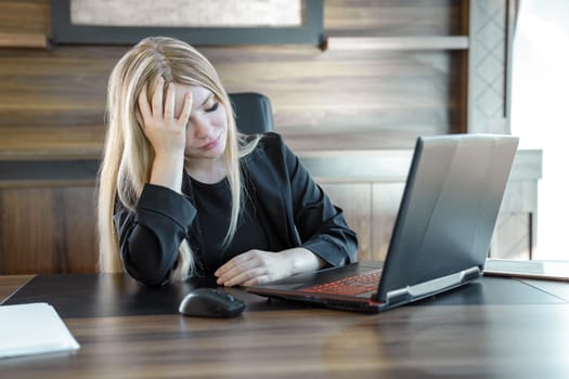 Distressed young business woman in black suit is sitting with eyes closed in front of laptop in office.