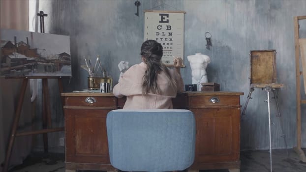Rear view of a brunette woman sitting at her desk and touching her hair. Woman working in the art workshop on grey walls background.
