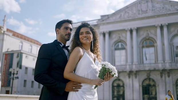 Elegant couple happily hugging. Action. Stylish couple in luxurious outfits hugging in city center. Newlyweds hug on background of ancient buildings on sunny summer day.