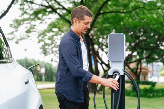 Man recharge EV electric vehicle's battery from EV charging station in outdoor green city park scenic. Eco friendly urban transport and commute with eco friendly EV car travel. Exalt