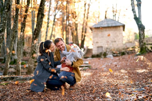 Mom crouches and hugs dad with a little girl on his knees in the park under falling leaves. High quality photo