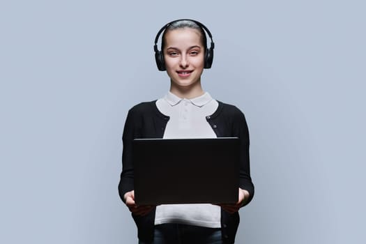 Teen girl student wearing headphones using laptop, looking at camera on grey studio background. Audio video internet technologies in education learning, online lessons, adolescence high school concept