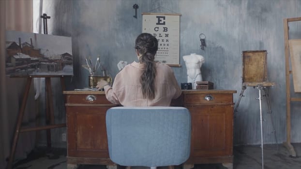 Businesswoman typing something on laptop and sitting at the desk, rear view. Back view of brunette woman sitting in the art workshop.