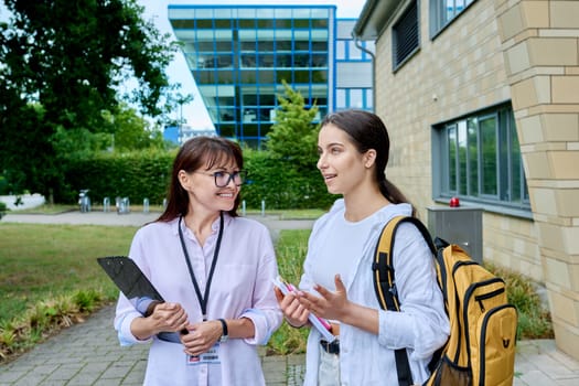 Teenage girl high school student with backpack talking to female teacher, mentor, coach, standing outdoors on educational building background. Adolescence, education, knowledge, communication