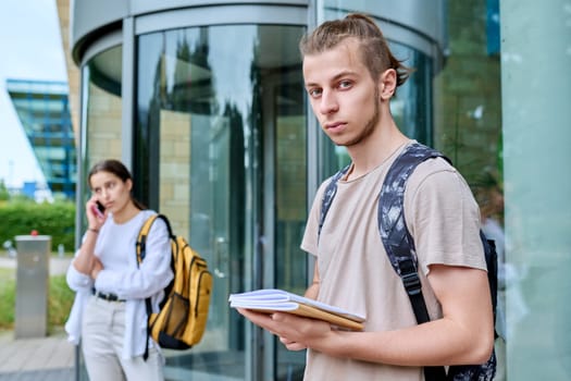 Hipster guy teenager student 18, 19 years old with backpack notebooks looking at camera, standing outdoor educational building. Youth, education, lifestyle concept