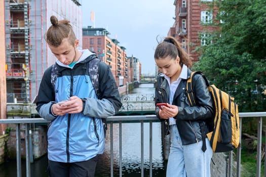 Teen friends guy and girl standing together holding smartphones looking at screen using mobile phones outdoor on city. Internet digital technology applications for leisure study communication