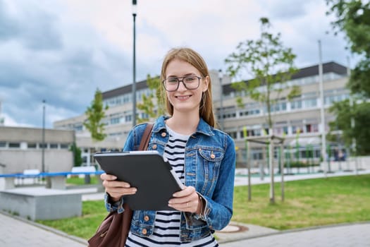 Girl student teenager outdoor near school building. Smiling teenage female with backpack digital tablet posing looking at camera. Adolescence, education, learning concept