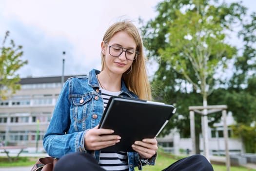 Girl student teenager outdoor near school building. Smiling teenage female with backpack, looking at screen of digital tablet. Adolescence, education, learning concept