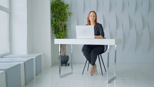 Woman in suit working in a modern office at the table. Action. White walls, green plant and a table with a laptop, minimalistic style