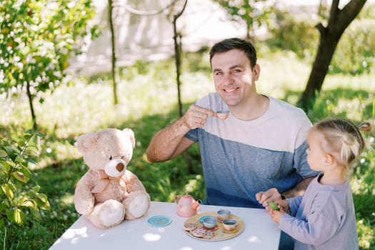 Little girl looking at smiling dad with toy cup at table with teddy bear. High quality photo